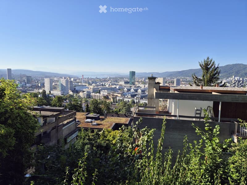 A modern building with a flat roof and a panoramic cityscape view in the background, surrounded by greenery.