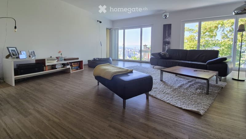 Modern living room with large windows offering a scenic view, featuring a dark leather sofa, wooden coffee table, blue ottoman, white shelf with picture frames and books, and a fluffy white rug.