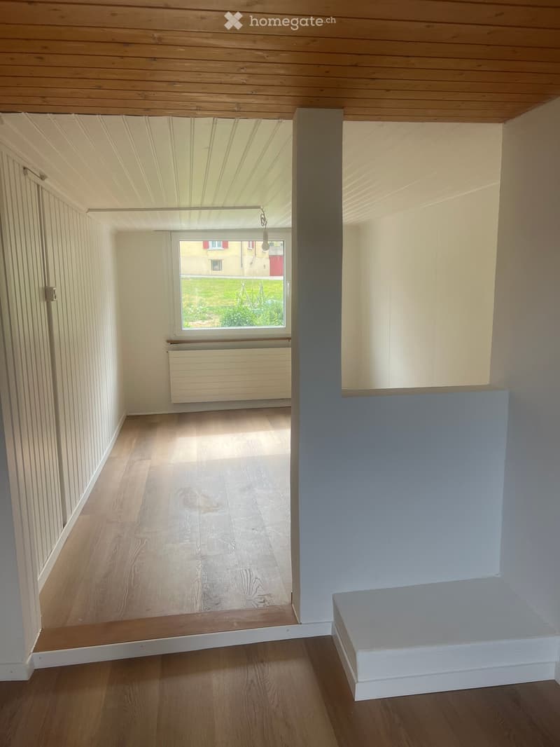 Empty room with wooden flooring, white walls, and a window showing a view of the outside. The room has a raised platform on one side and a radiator under the window.