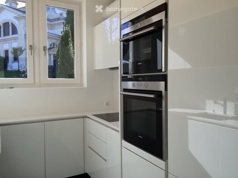 Modern kitchen with white cabinets, built-in oven, sink, and a view of a building and tree outside the window.