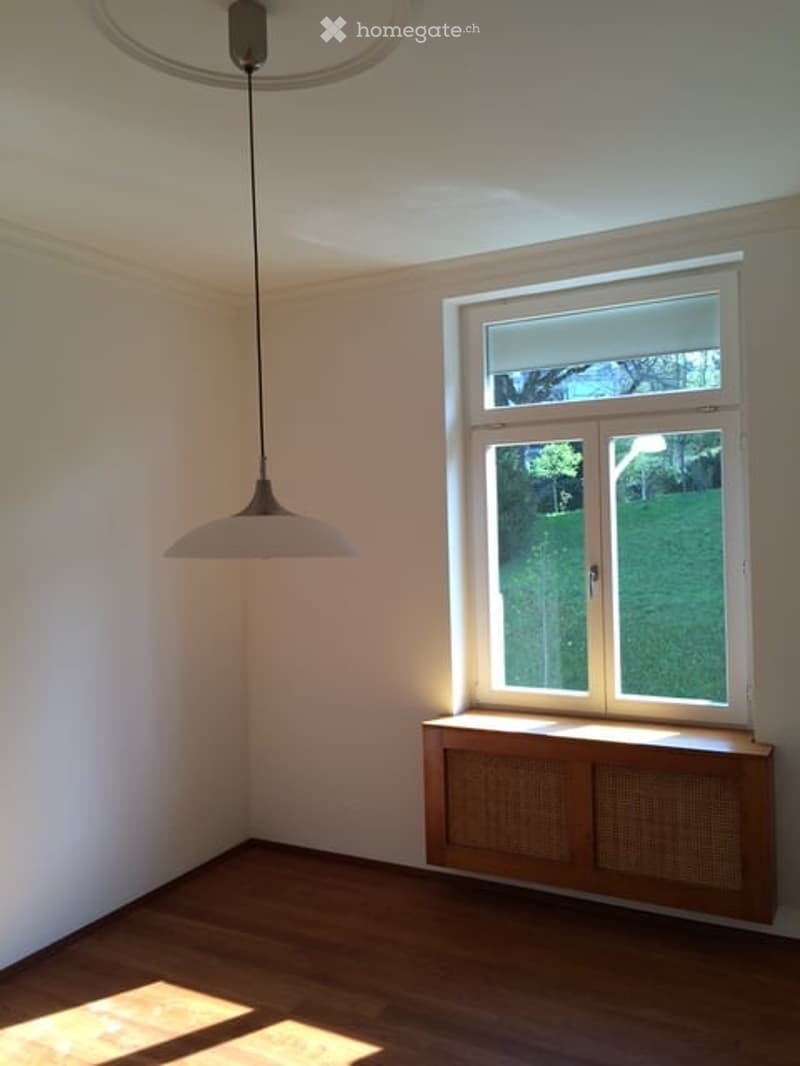 Empty room with wooden flooring, white walls, a large window with a view of a garden, and a hanging pendant light fixture.