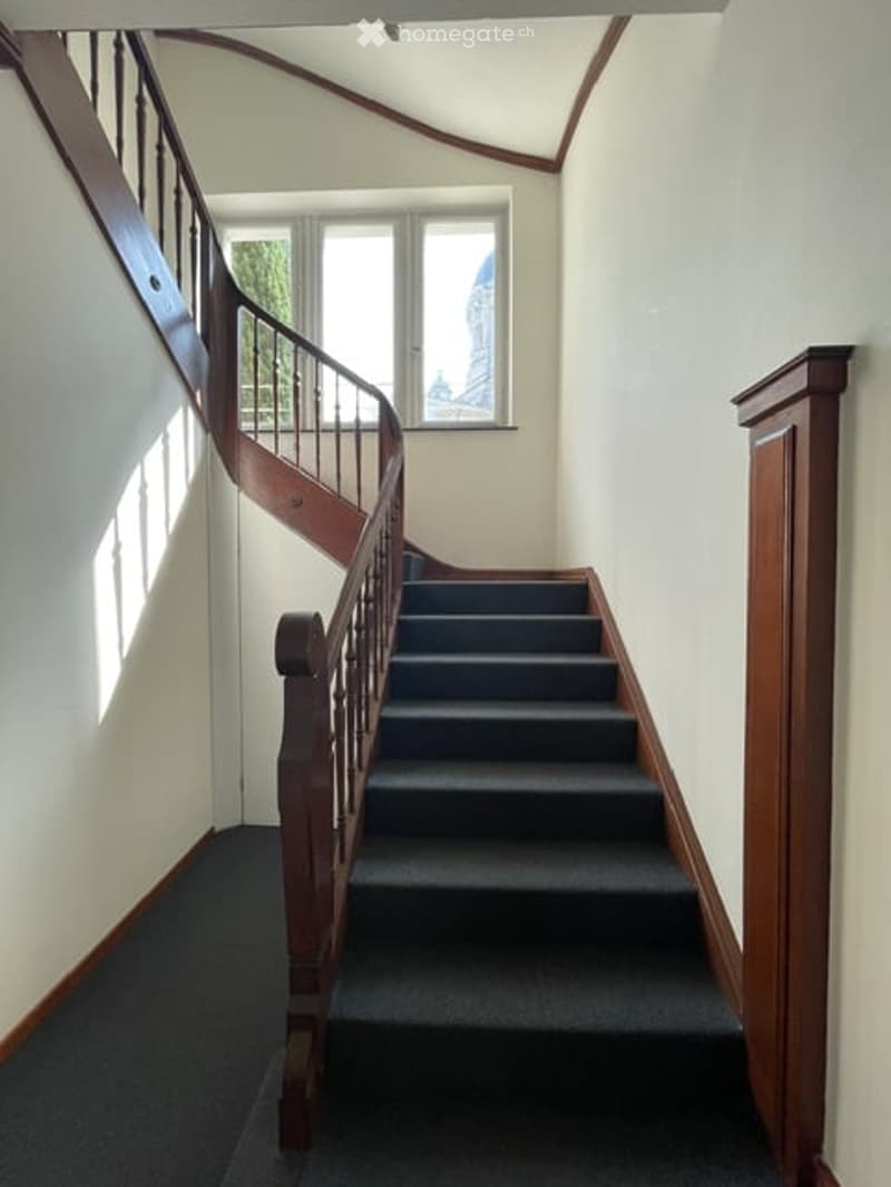 Spacious hallway with wooden staircase, grey carpeted steps, and white walls. Natural light from a window illuminates the area.