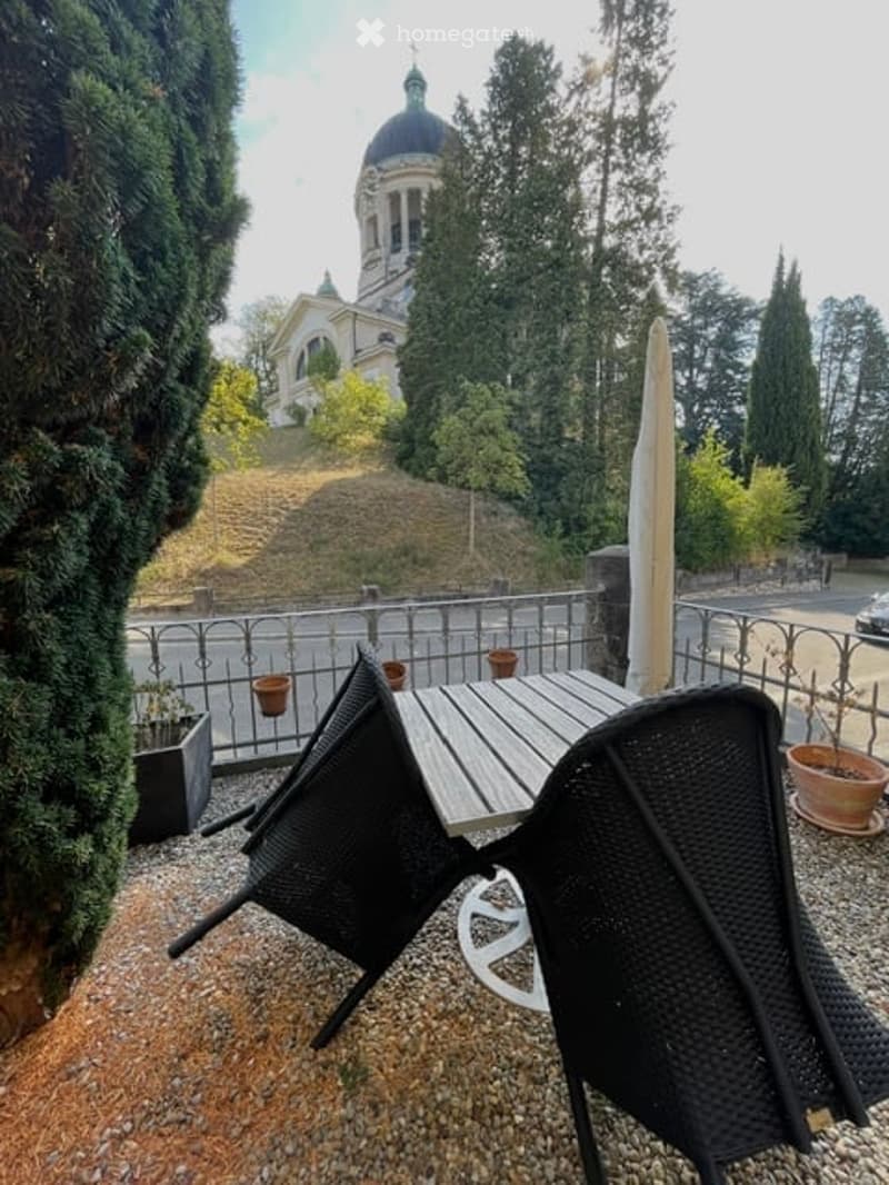 Outdoor patio with a wooden table, black chairs, umbrella, and potted plants, overlooking a scenic view of a domed building and greenery.