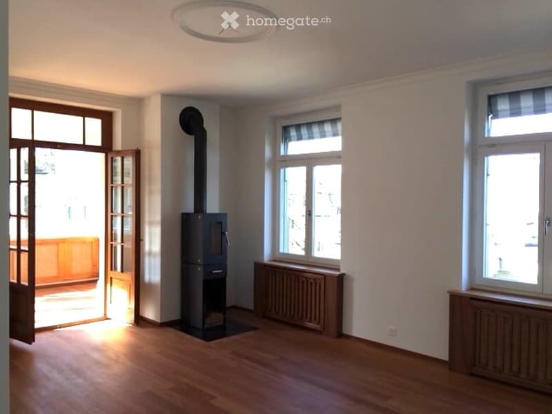 Empty room with wooden flooring, white walls, large windows, and a wood-burning stove in the corner.