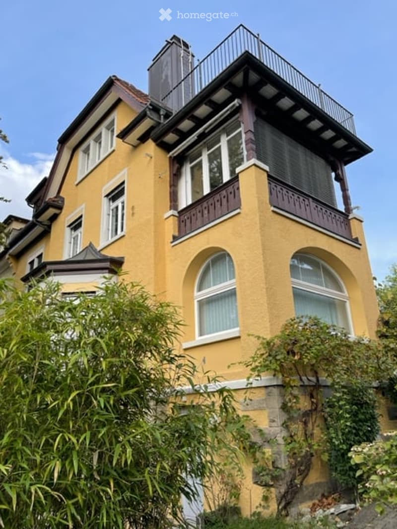 A multi-story yellow building with white-framed windows, a prominent balcony on the upper floor, and decorative wooden railings.