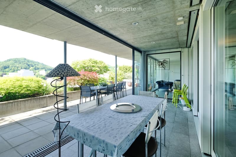 Modern balcony with a dining table, chairs, and a decorative lamp, overlooking a landscaped garden and hills.