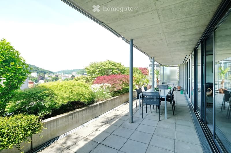 Modern balcony with tiled floor, glass walls, and outdoor dining set, overlooking a landscaped garden and cityscape.