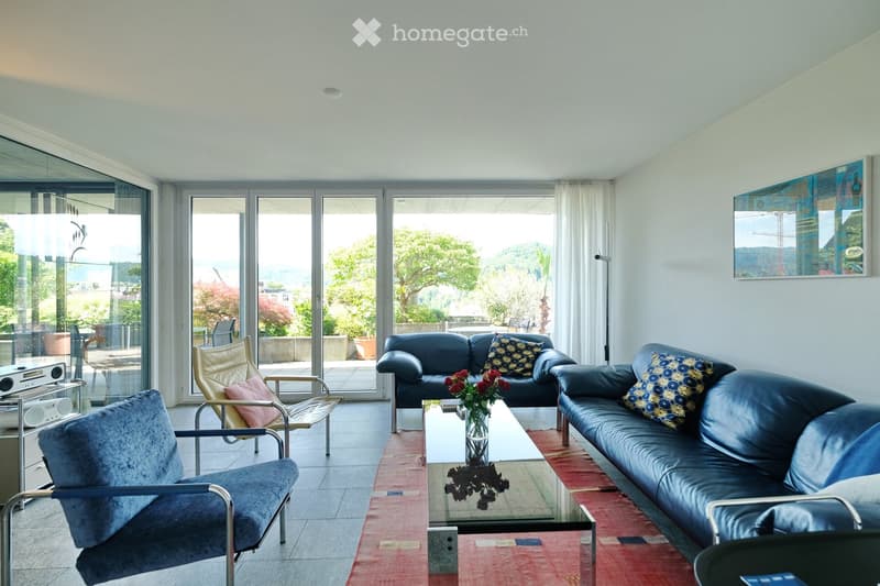 Modern living room with large glass doors offering a scenic view, featuring a dark blue leather sofa, glass coffee table with a flower arrangement, and several chairs including a blue velvet chair and a gold-framed chair.