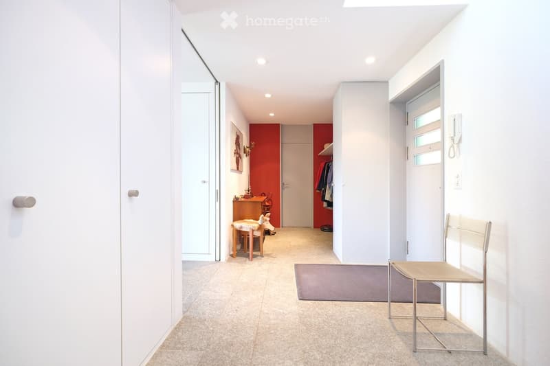 Modern hallway with white walls, tiled floor, recessed lighting, and a small seating area with a chair and a small table.
