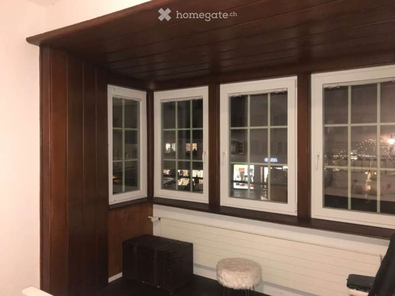 A small, cozy room with large windows, wooden paneling, a radiator, and a small stool.