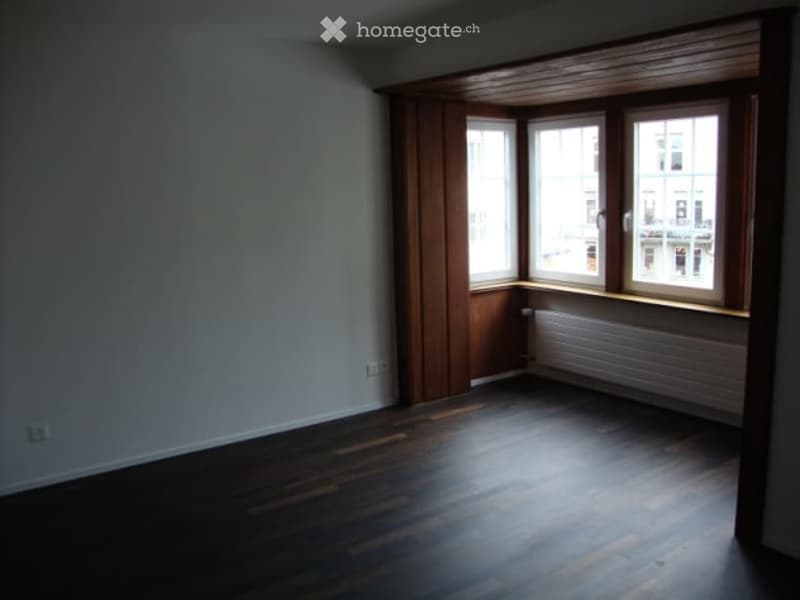 Empty room with wooden flooring, white walls, and large windows with wooden frames. Radiator visible under the windows.