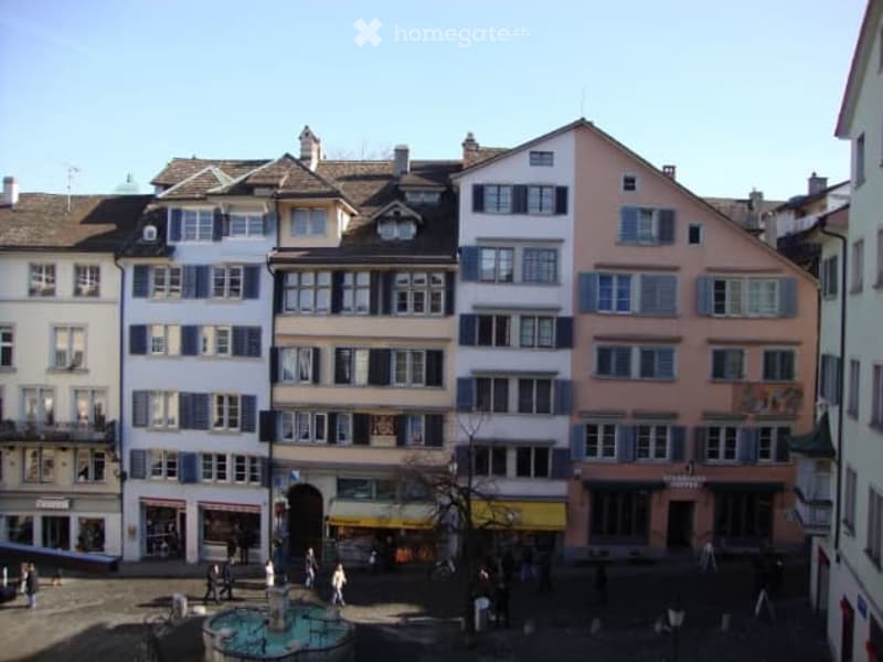 A row of traditional European townhouses with varied facades, featuring multiple stories, windows, and a central square with a fountain and people walking.