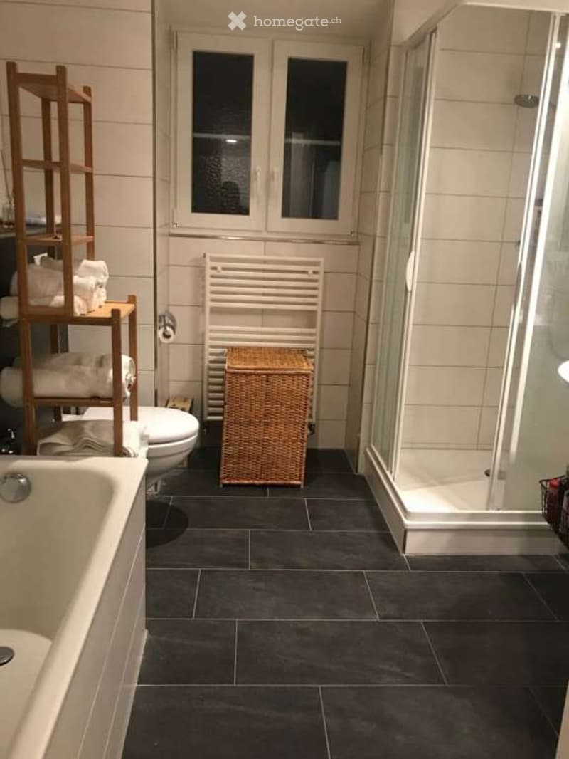 Modern bathroom with dark tiled floor, white tiled walls, bathtub, toilet, shower, towel rack, wicker basket, and wooden shelf holding towels.