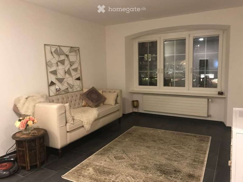 Modern living room with a white tufted sofa, decorative pillows, a fur throw, a patterned rug, a side table with a flower vase, and a geometric wall mirror. Large windows provide natural light.
