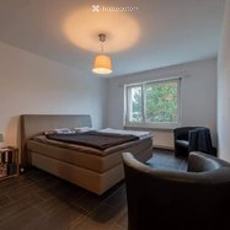 Bright bedroom with wooden flooring, large windows, a bed with dark bedding, a nightstand, a chair, and a lamp.