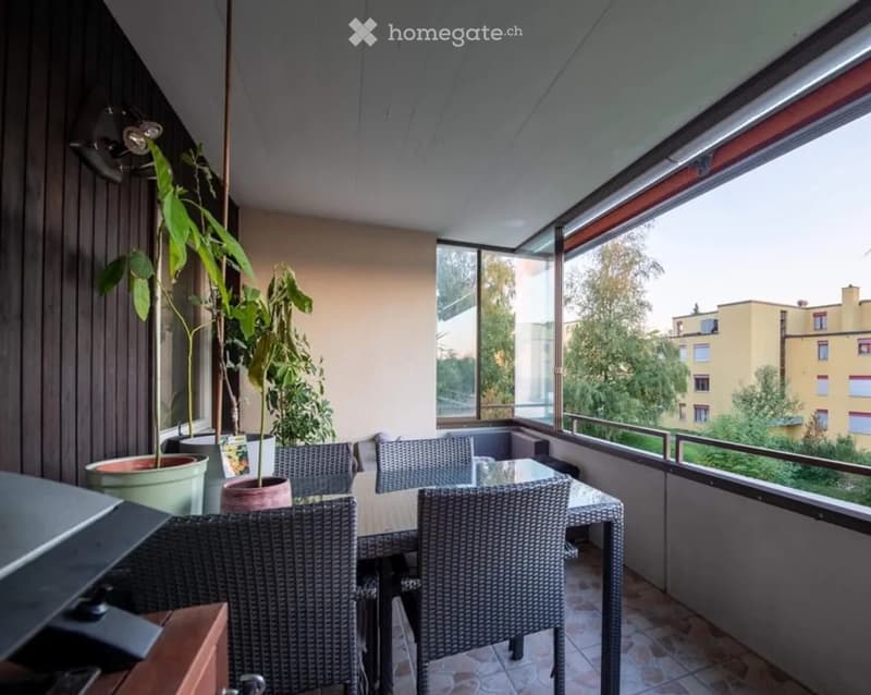 Balcony with a glass table, wicker chairs, and potted plants, offering a view of the city and greenery.