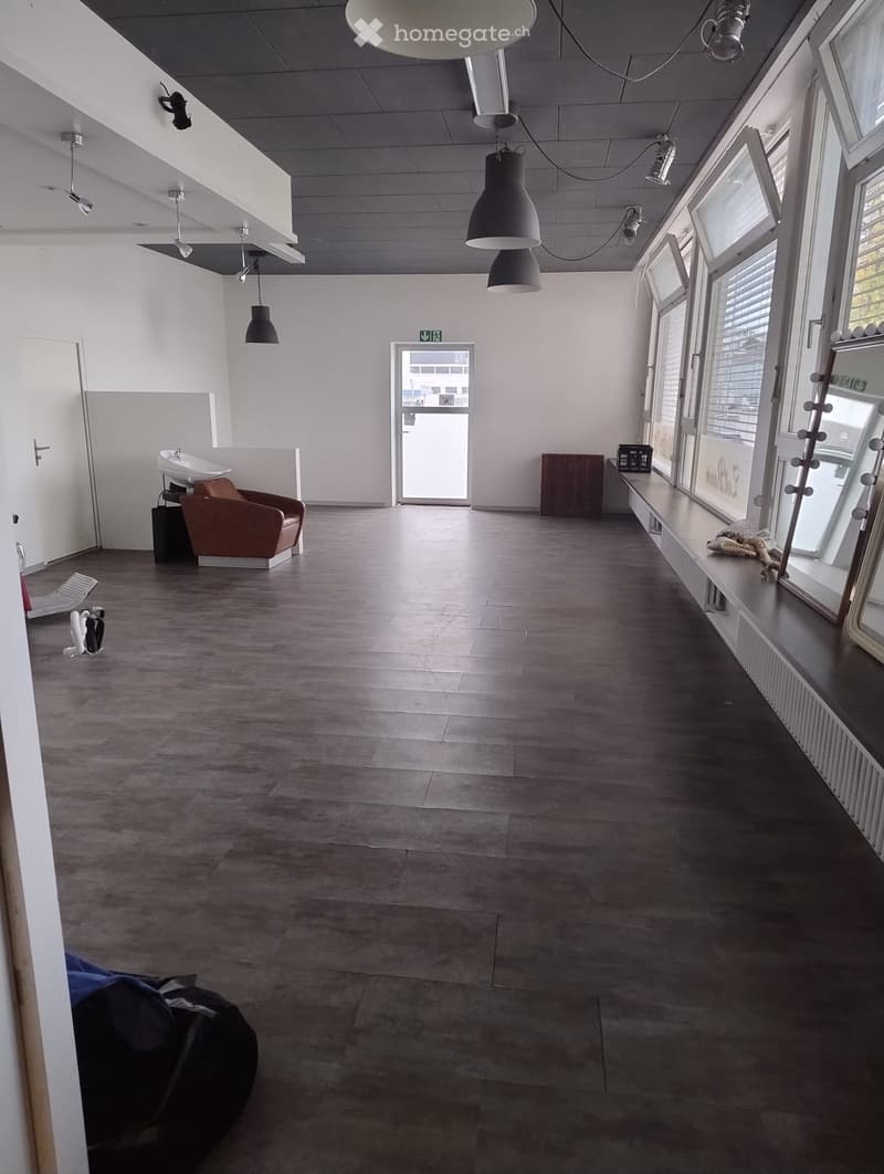 Modern, minimalist room with dark tiled flooring, large windows, a single chair, a sink, and a mirror. The space is well-lit with several hanging lamps and has a clean, contemporary design.