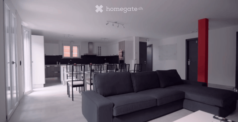 Modern living and dining area with a grey sectional sofa, dining table with chairs, and an open kitchen with white cabinets and stainless steel appliances.