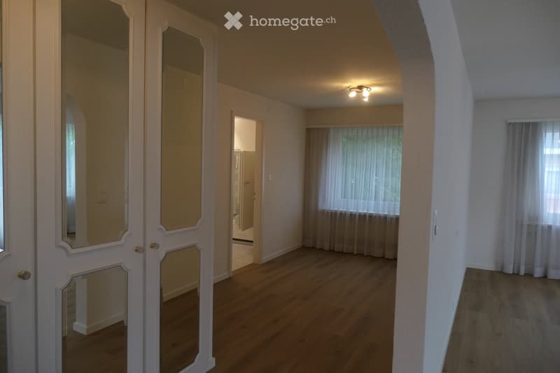 Empty hallway with wooden flooring, white walls, and large windows with sheer curtains. The hallway leads to a room with a visible bathroom area.