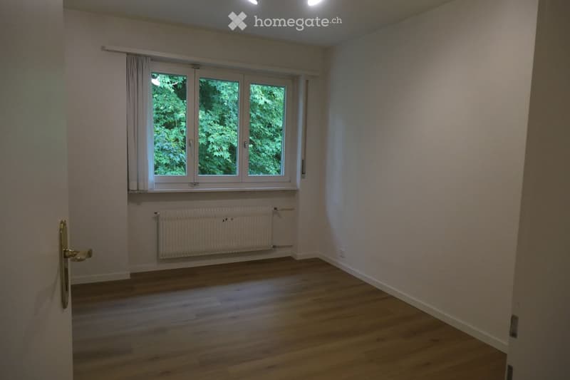 Empty room with wooden flooring, white walls, large window with white frame, radiator, and a door with a golden handle.