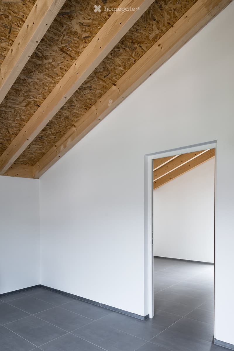Empty room with exposed wooden beams and insulation in the ceiling, grey tiled floor, white walls, and a doorway leading to another room.