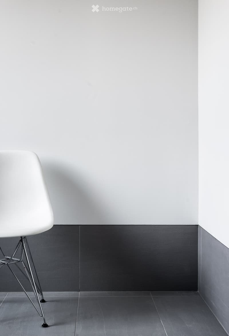 Empty room with white walls, dark grey lower wall paneling, and grey tiled floor. A single white chair is visible in the corner.