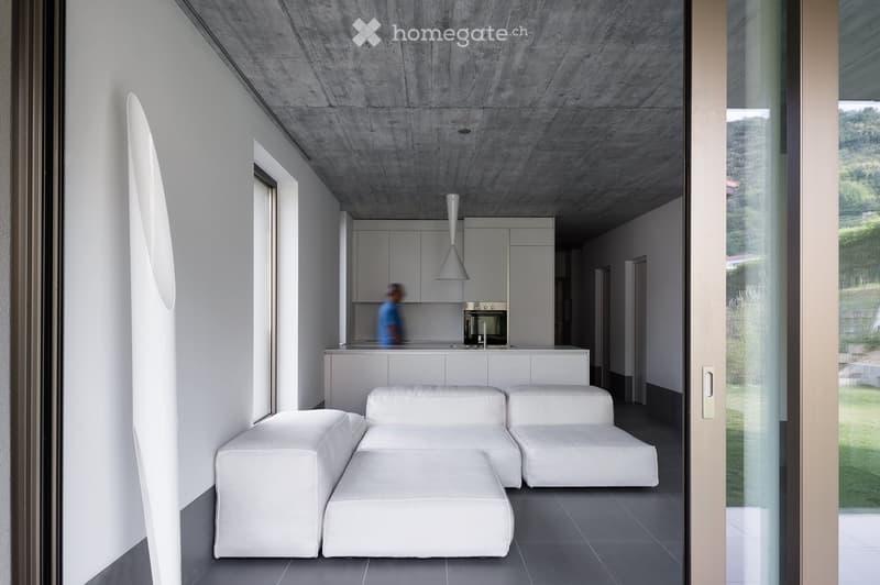 Modern living space with white sectional sofa, ottoman, and open kitchen area featuring white cabinets and stainless steel appliances.