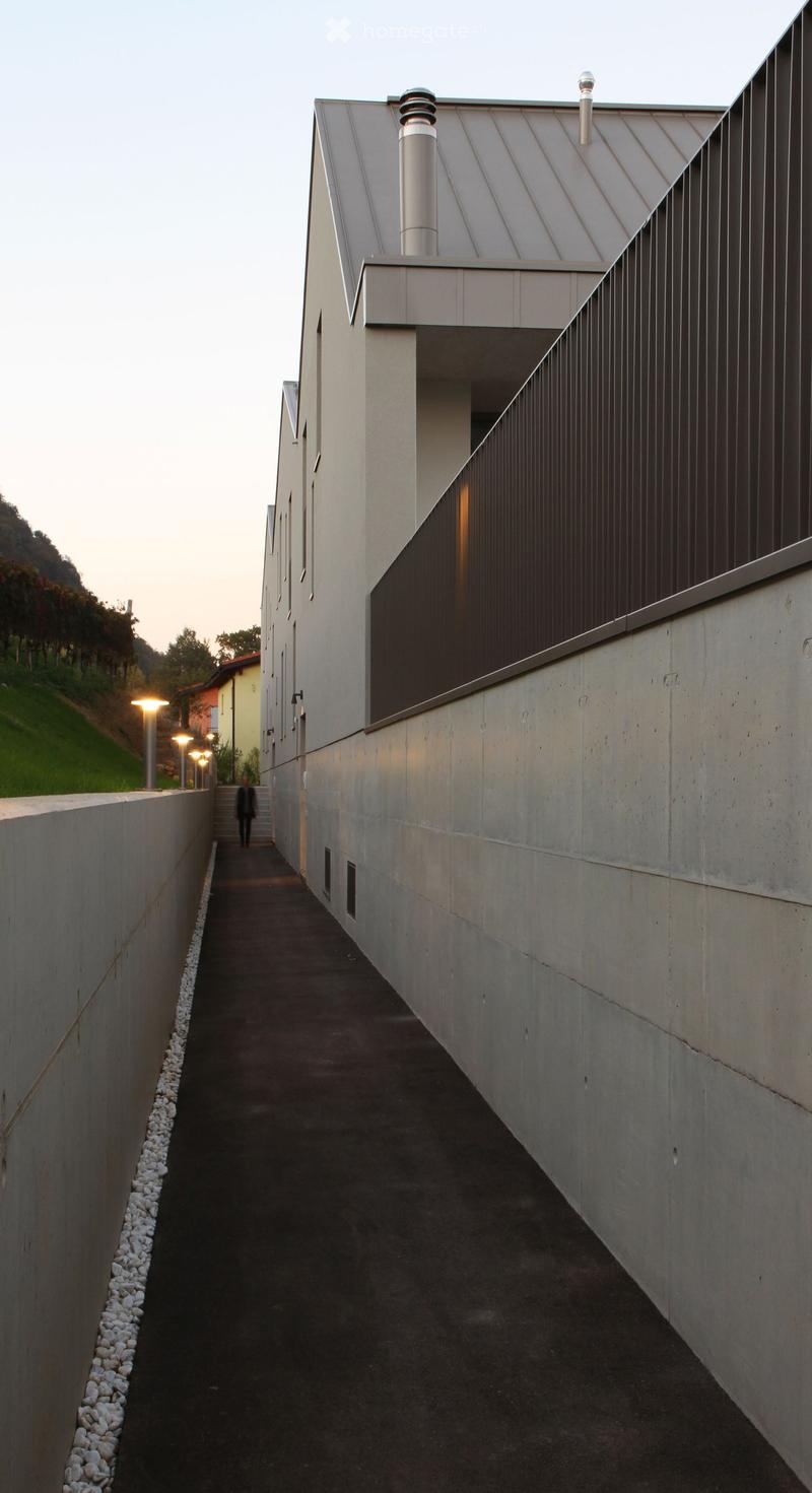 A modern, narrow concrete corridor with a raised ground floor, flanked by tall concrete walls, leading to a modern building with a metallic roof and chimney.