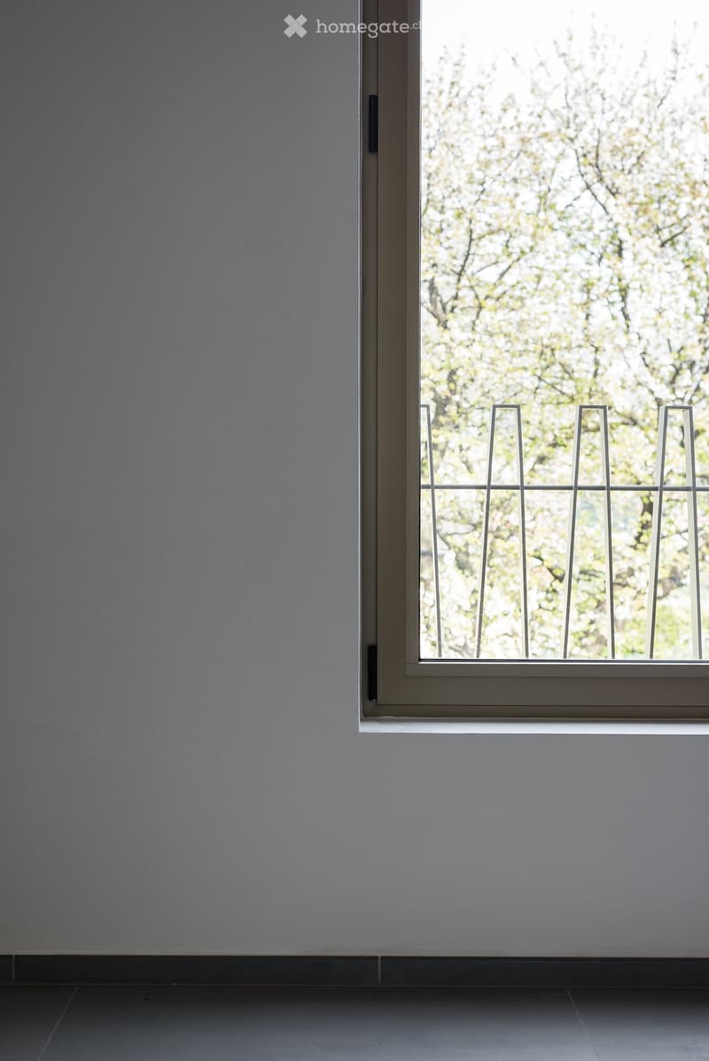 Empty room with grey walls and a large window with a metal security grate, showing a view of blooming trees outside.