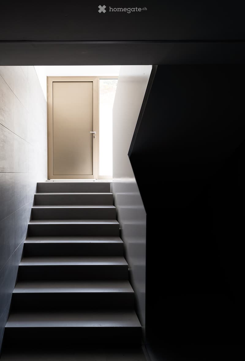 Modern, minimalist staircase with grey steps and a sleek, contemporary design. The walls are white and grey, and there is a door at the top of the stairs.