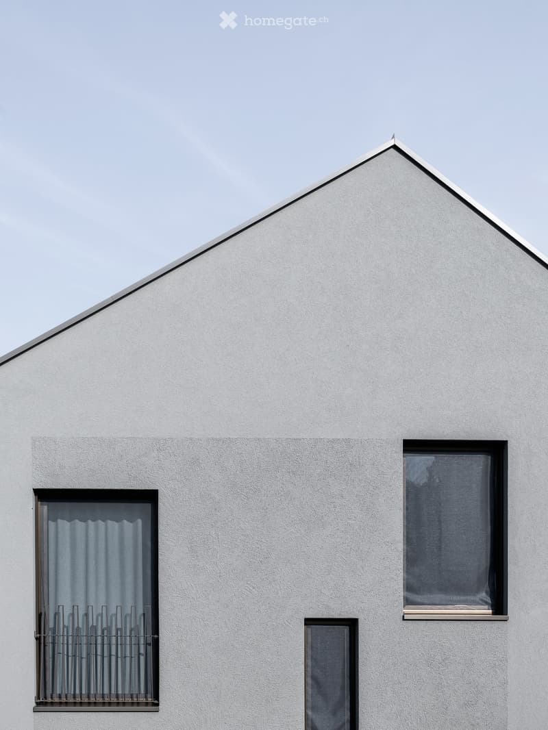 Modern white house with a triangular roof and three windows, one with a metal security grate and curtains.