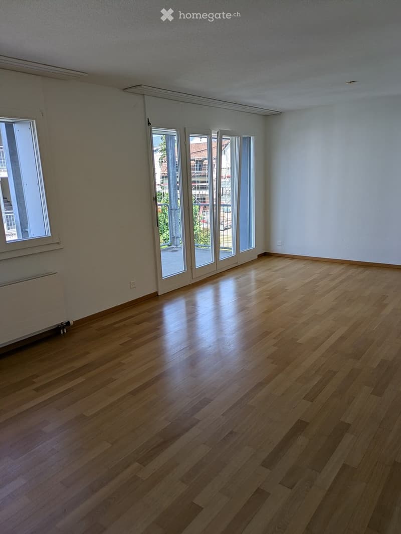 Empty room with wooden flooring, large windows, and sliding glass doors leading to a balcony.