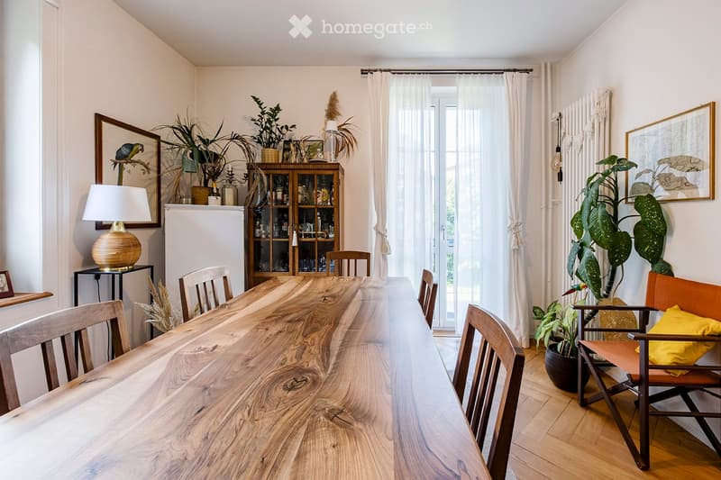 Bright dining room with a long wooden dining table, multiple chairs, a glass-fronted cabinet, a lamp, and several potted plants. Large windows with white curtains let in natural light.