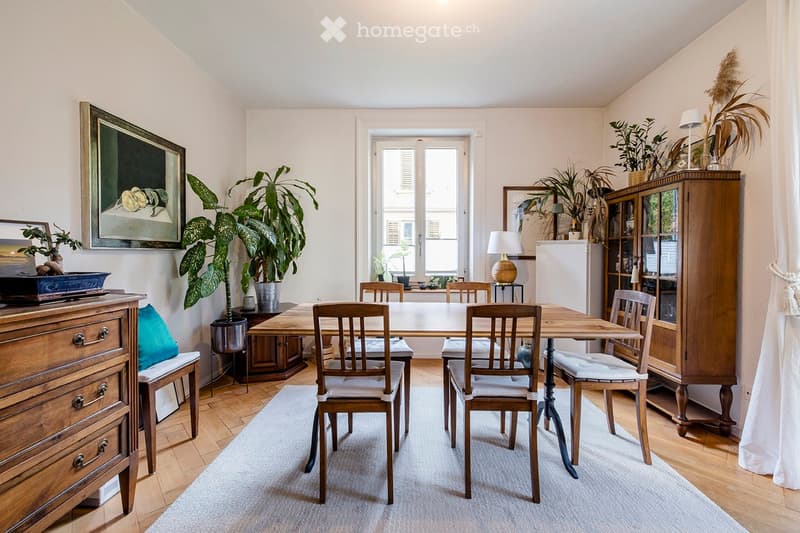 Cozy dining room with a wooden dining table and chairs, a wooden cabinet, a lamp, and several potted plants. The room features light-colored walls, wooden flooring, and a large window with shutters.