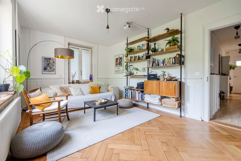 Modern living room with white walls, wooden flooring, and large windows. Features a white sofa with colorful cushions, a wooden coffee table, a wicker chair, a floor lamp, and a bookshelf filled with books and decorative items.