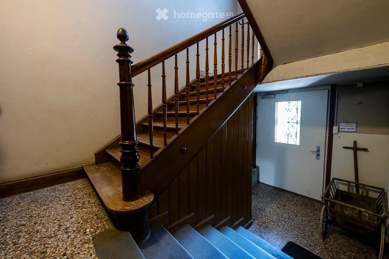Wooden staircase with handrail and balusters, leading to a doorway with frosted glass window. A small wooden cart is visible on the right side of the hallway.
