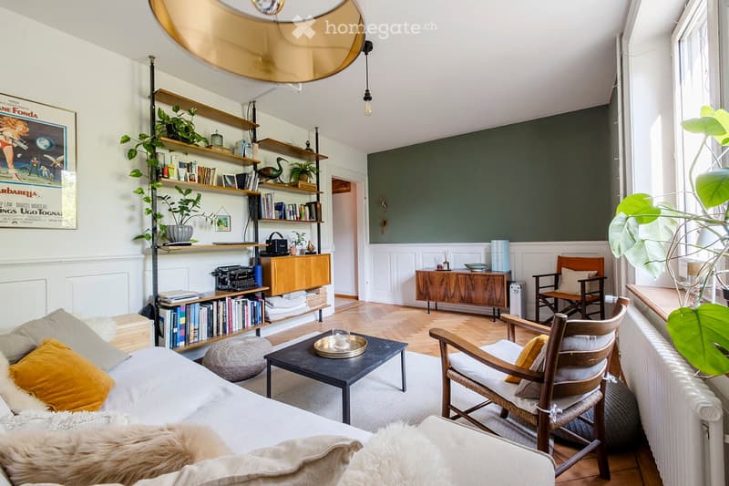 A modern living room with a white sofa, wooden coffee table, wooden chairs, shelves filled with books and decorative items, and large windows letting in natural light.