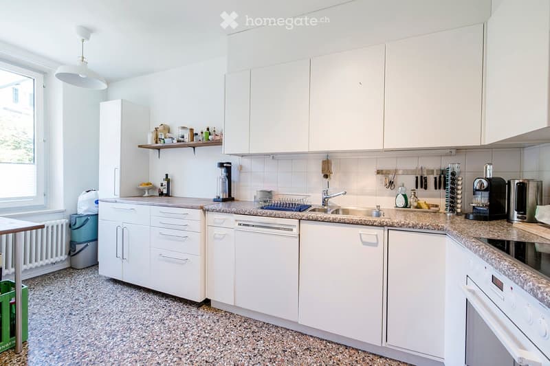 Bright kitchen with white cabinets, granite countertops, and modern appliances including a dishwasher, oven, and coffee maker. Shelves hold various kitchen items and a fruit bowl.