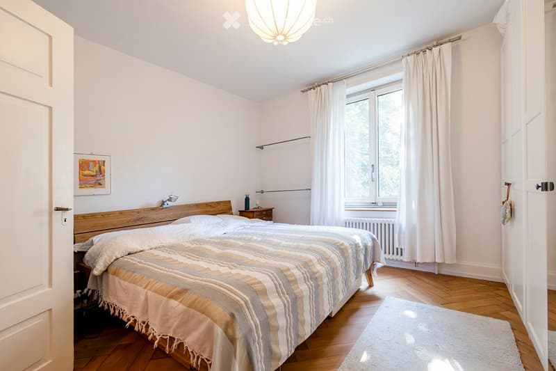 Cozy bedroom with wooden bed, nightstand, lamp, and large window with curtains. Light-colored bedding and a small rug on the floor.