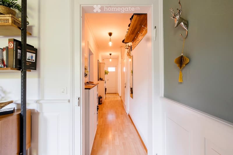 A well-lit hallway with wooden flooring, white walls, and several doors leading to different rooms. A coat rack with hooks is mounted on the wall, and a decorative deer head is visible on the right side.