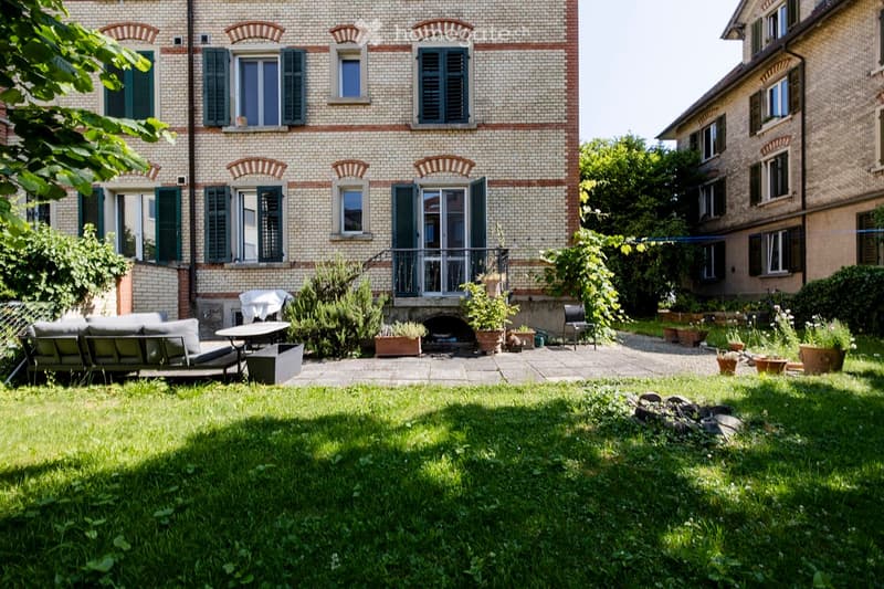 Garden with green grass, a sofa, a table, a chair, and several potted plants. The background features a brick building with multiple windows and shutters.