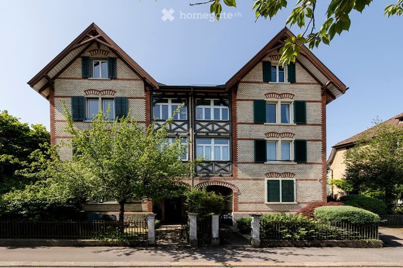 Two-story brick house with green shutters, decorative wooden beams, and a fenced garden.