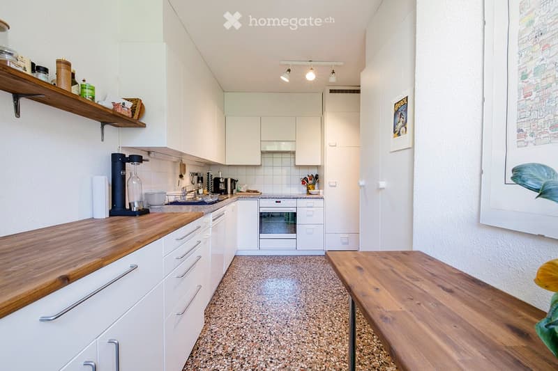 Modern kitchen with white cabinets, wooden countertops, built-in oven, and various appliances. Features a window with a view and a small wooden table.