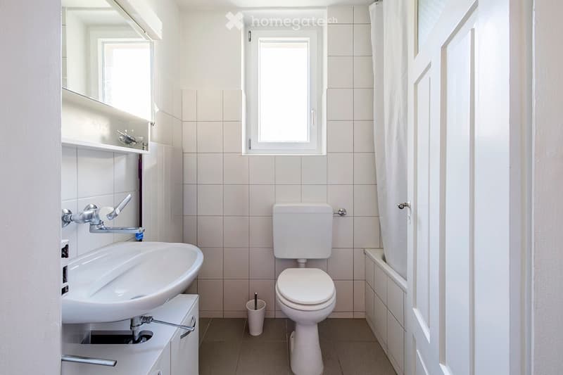 Modern bathroom with white tiles, a sink, toilet, bathtub, and a mirror above the sink. The space is bright with a window and a small shelf holding a few items.