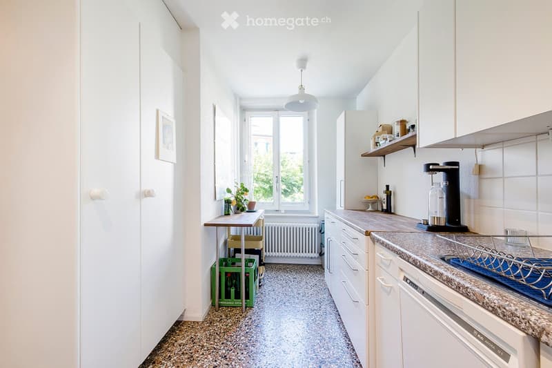 Bright, modern kitchen with white cabinets, granite countertops, and a small table with a plant. Features include a refrigerator, dishwasher, coffee maker, and shelves with various items.