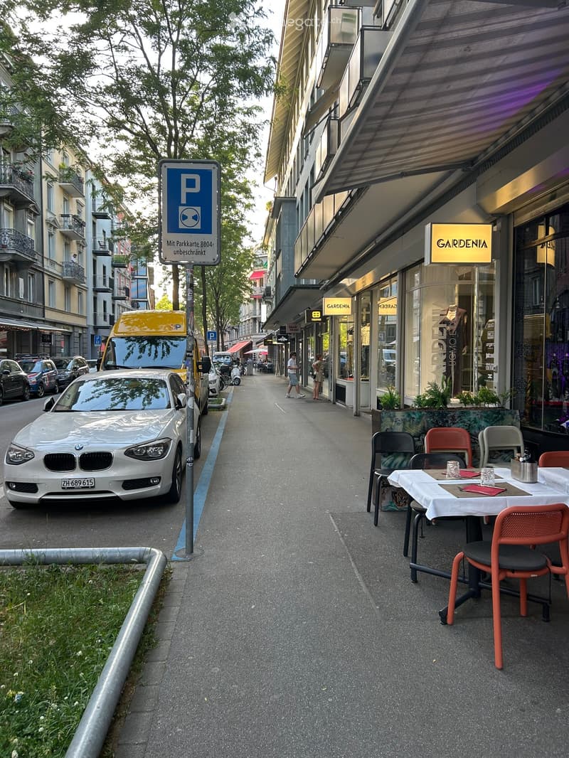 A narrow pedestrian street lined with outdoor seating from a café, with a few parked cars and a parking sign visible.