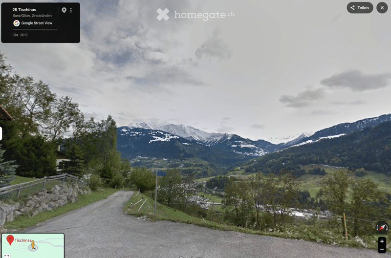 A scenic outdoor view showing a winding road leading through a lush green valley with scattered houses, surrounded by snow-capped mountains under a cloudy sky.