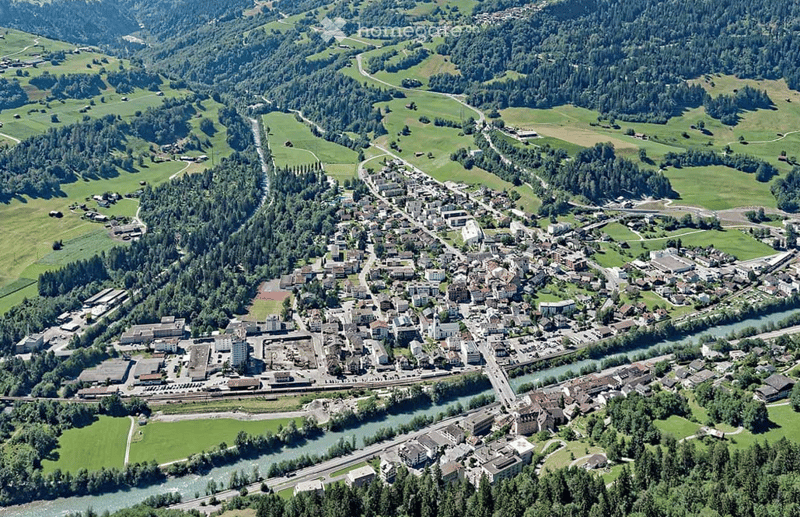 Aerial view of a town nestled in a valley surrounded by green hills and forests. The town features a mix of residential and commercial buildings, roads, and a river running through it.