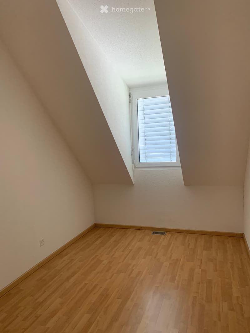 Empty attic room with wooden flooring, slanted ceiling, and a single window with blinds.