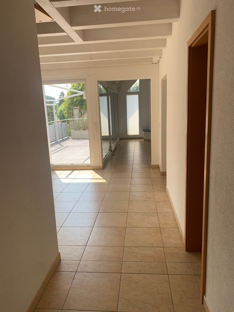 Bright hallway with tiled flooring, white walls, and large glass doors leading to a balcony area with outdoor space and greenery visible.